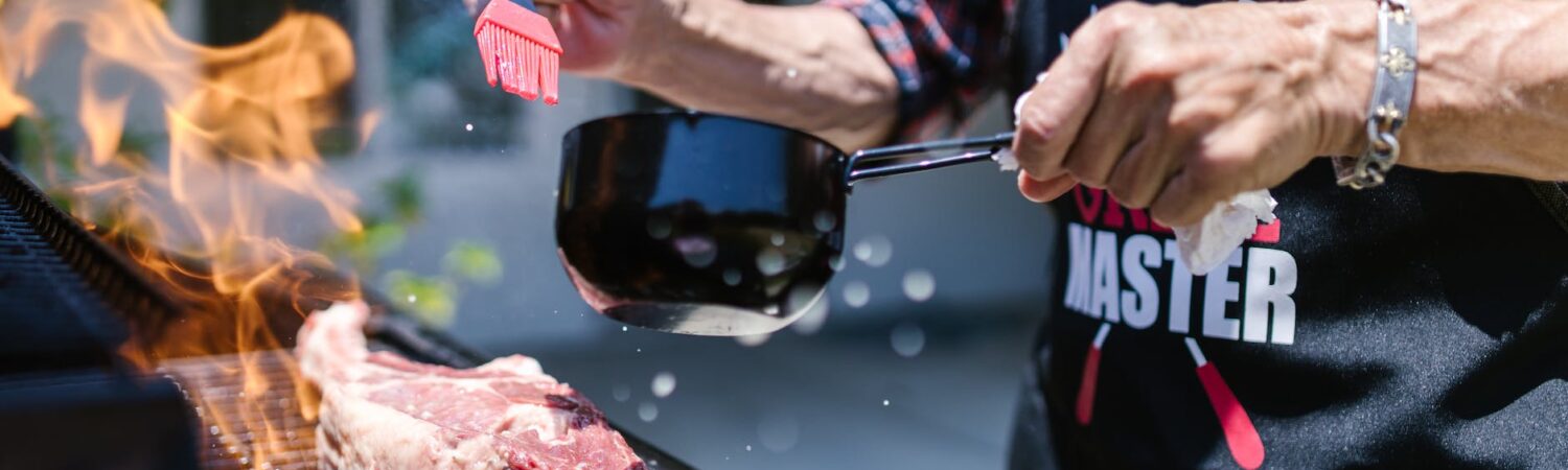 man in black apron putting sauce on meat while grilling