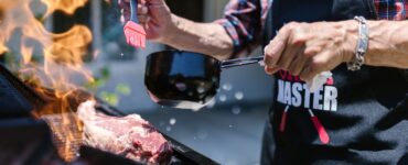 man in black apron putting sauce on meat while grilling