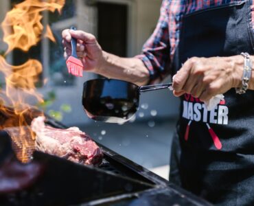 man in black apron putting sauce on meat while grilling