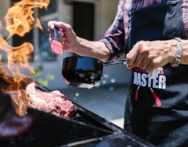 man in black apron putting sauce on meat while grilling